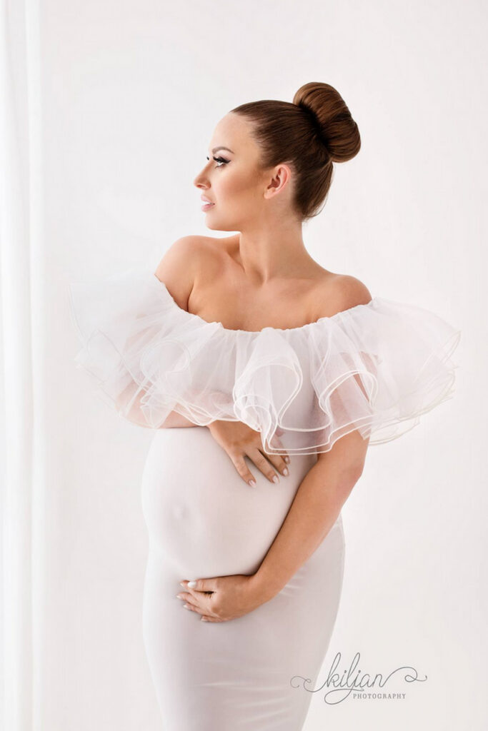 Pregnant woman in elegant off shoulder gown with voluminous tulle neckline, studio portrait