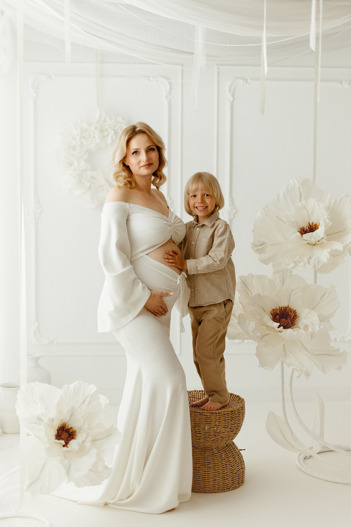 Pregnant woman in elegant white maternity dress posing with her son in a bright studio, luxury family maternity photoshoot outfit by Kamaprops.