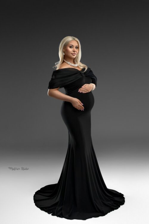 Pregnant woman wearing a black off-shoulder maternity gown with fitted silhouette and elegant styling against a studio background