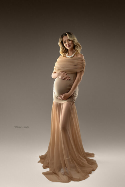 Pregnant woman wearing a beige tulle maternity gown with off-shoulder draped top standing against a neutral studio background