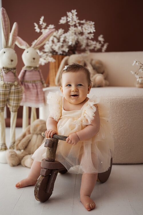 Baby girl wearing a beige tulle romper with pearl details sitting on a wooden ride-on toy in a styled studio setup