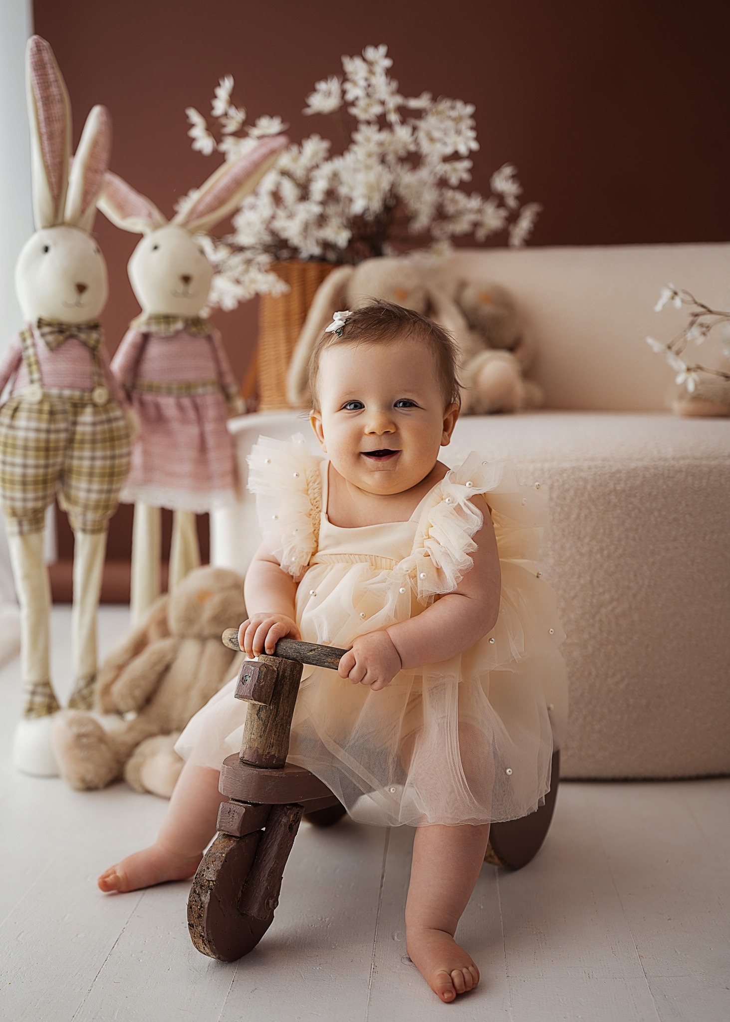 Baby girl wearing a beige tulle romper with pearl details sitting on a wooden ride-on toy in a styled studio setup