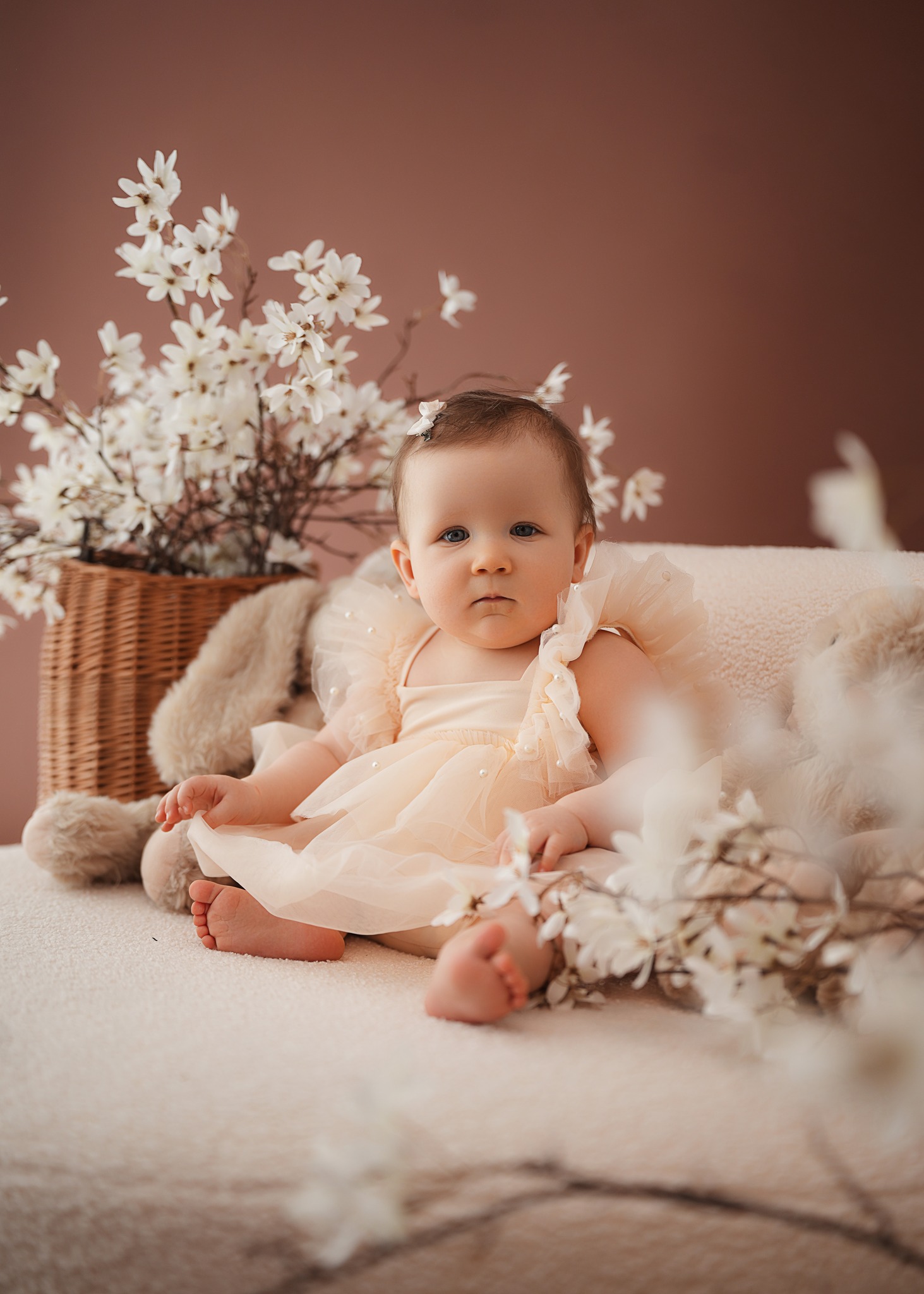 Baby in a soft tulle romper with pearls sitting on a neutral sofa with floral decorations in the background