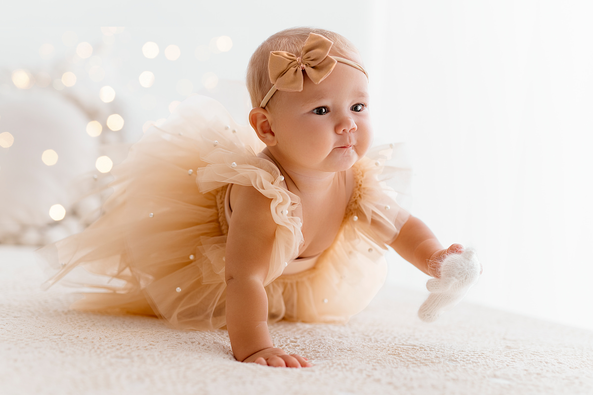 Baby girl crawling on a soft blanket wearing a beige tulle romper with pearl details and a bow headband