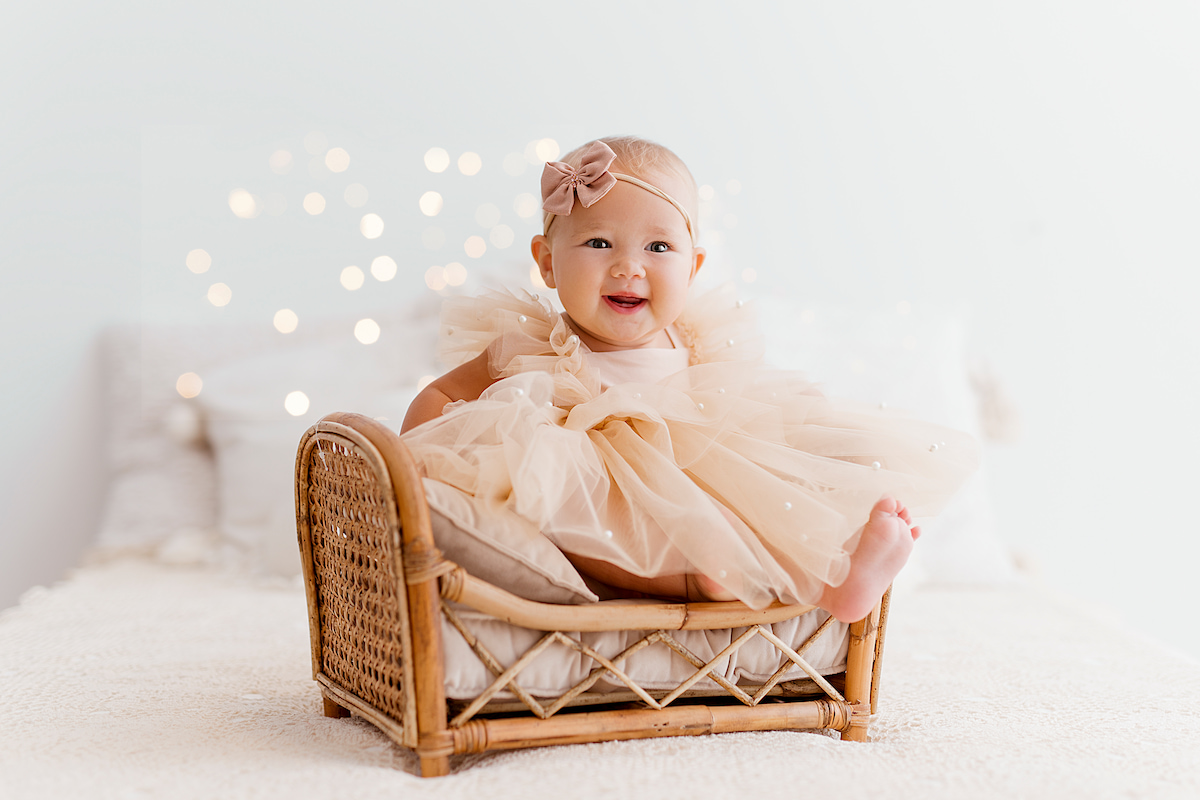 Smiling baby sitting in a small wicker bed wearing a tulle romper with pearls in a bright minimal studio setting