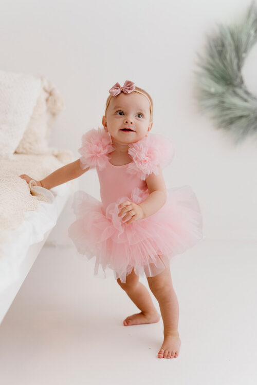 Baby girl wearing a pink tulle romper with floral shoulder details standing next to a bed in a bright studio